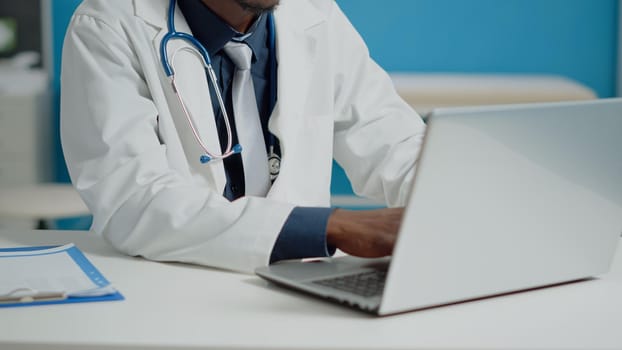 Close up of doctor typing on laptop keyboard in medical cabinet at healthcare facility. Portrait of medic sitting at desk with documents using device with technology for consultations