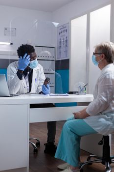 Medic of african american ethnicity talking to old patient and holding bottle of pills for healing treatment. Doctor and senior woman doing consultation wearing face masks during pandemic