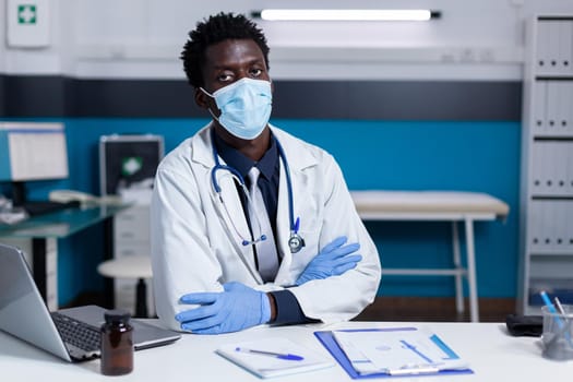 Portrait of african american medic looking at camera wearing gloves and face mask for protection. Black doctor with stethoscope having bottle of pills, laptop and documents on desk