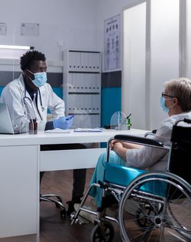 Caucasian patient with disability receiving consultation from african american doctor in medical cabinet. Invalid elder woman sitting in wheelchair for appointment during pandemic