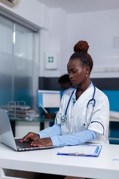 African american person with medic job using laptop at desk in medical office. Black doctor sitting with technology and healthcare equipment while nurse looking at computer in background