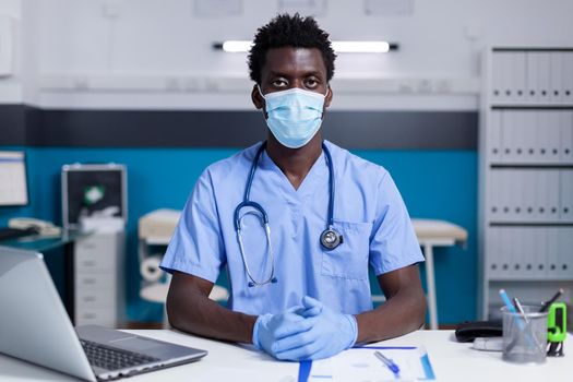 Portrait of african american man with nurse occupation sitting at desk with laptop, documents and medical tools. Black person working in medical cabinet having stethoscope and face mask