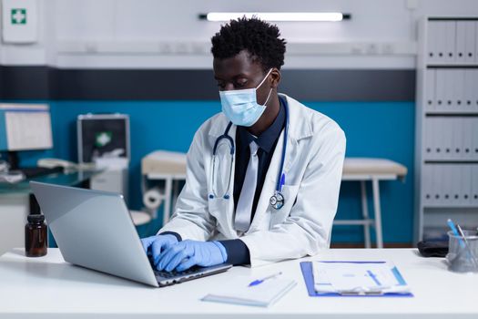 Black man with doctor occupation using laptop on desk for medicine prescription in medical office. African american healthcare specialist typing on keyboard while wearing face mask