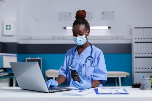 Portrait of african american nurse typing on laptop keyboard while looking at bottle of pills for medical advice. Black woman assistant wearing uniform, gloves and face mask sitting at desk