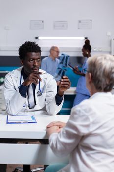 Medic of african american ethnicity holding x ray scan explaining disease to elder woman in cabinet. Black man and old patient doing checkup while nurse and senior man talking in background