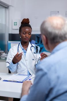 African ethnicity woman with doctor job looking at x ray scan in hand while discussing with senior ill patient at healthcare clinic. Black medic holding radiography for old man with disease