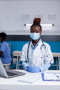 African american doctor sitting at desk with laptop computer in healthcare office. Black woman with medic profession wearing face mask and stethoscope for examination at medical clinic