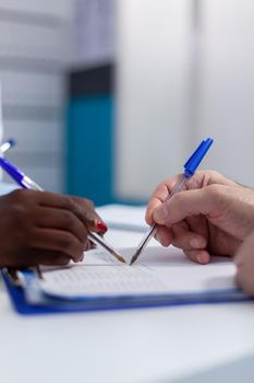 Close up of hands holding pen on document files for signature at healthcare office desk. Caucasian senior man signing appointment papers for checkup with african american doctor