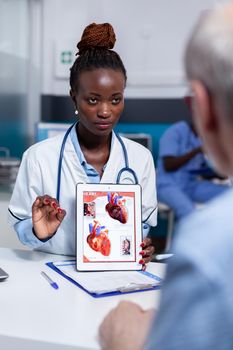 Black woman with doctor profession showing organs illustration on digital tablet display sitting at desk. African american medic explaining diagnosis to elderly sick patient in cabinet