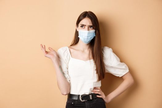 Covid-19 and lifestyle concept. Confused girl in face mask looking at upper left corner with troubled face, standing on beige background.