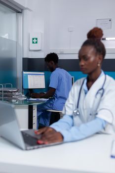 Portrait of african american woman with doctor profession sitting at desk while using laptop and technology in healthcare office. Black medic with modern device and man nurse in background