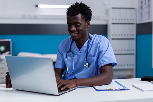 Black young man working as nurse at medical clinic wearing uniform and stethoscope. African american person sitting at desk while using modern laptop, typing on keyboard in cabinet