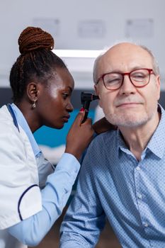 Otologist of african ethnicity consulting elder patient using professional otoscope in healthcare office. Black doctor with metal tool for ear checkup and stethoscope checking on senior man