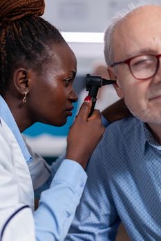 Close up of black doctor using otoscope for ear checkup examination on old patient in cabinet. African american otologist holding medical steel instrument for healthcare consultation