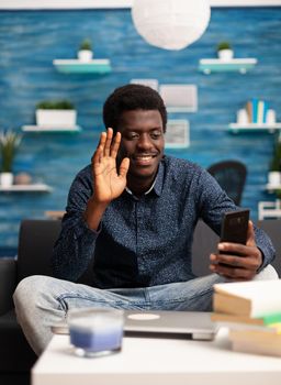 Black man talking on online video call conference in bright living room, using smartphone technology. African american internet online user working from home for office project planning