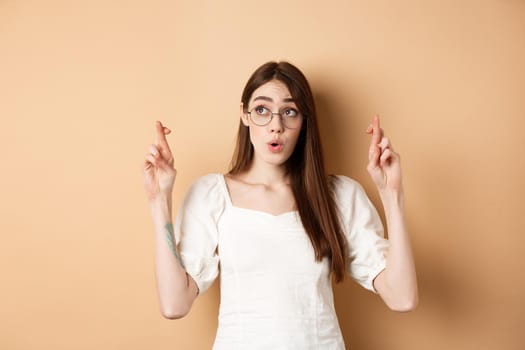 Excited girl hope dream come true, making wish with fingers crossed, looking aside and praying, standing on beige background.