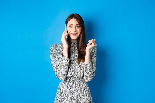 Excited woman receive good news on mobile phone, smiling happy, talking on smartphone, standing on blue background.
