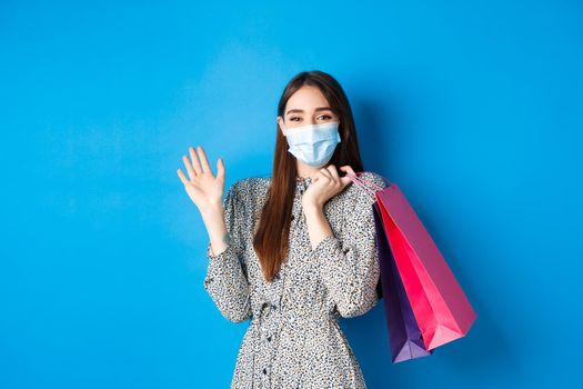 Covid-19, pandemic and lifestyle concept. Friendly cute woman wear medical mask in shopping mall, waving hand and holding bags, say hello, standing on blue background.