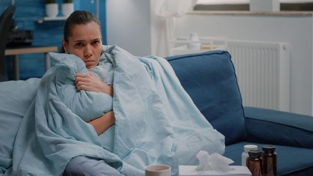 Unhappy woman shivering because of cold and flu, using blanket and pillow for warmth. Sick adult with virus disease looking at camera while waiting on treatment to cure illness.