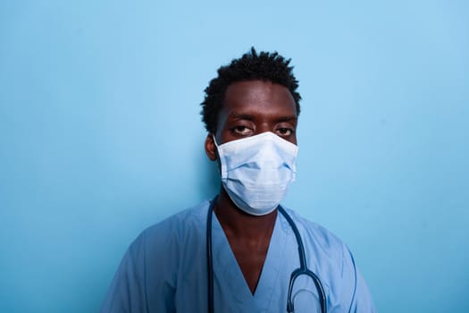 Portrait of black medical assistant wearing face mask and stethoscope over isolated background. African american nurse with blue uniform and protection against coronavirus pandemic.