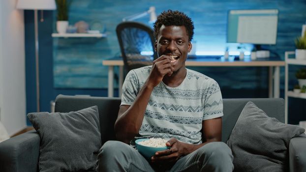 POV of adult watching comedy on television with popcorn. Person enjoying fun movie on TV, having snack and looking at camera for entertainment. Man with film on screen laughing