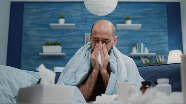 Close up of man with cold and flu blowing runny nose using tissue. Sick adult with virus symptoms and disease wrapped in blanket having medicaments against infection and tissues.