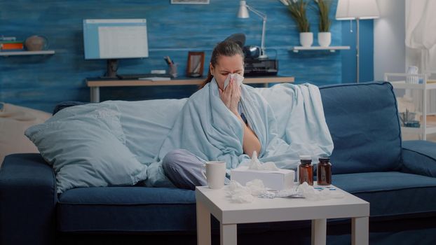 Unhappy woman with flu using tissue to blow runny nose. Adult with disease and virus symptoms wrapped in blanket and drinking cup of tea to cure sickness. Ill person blowing nose