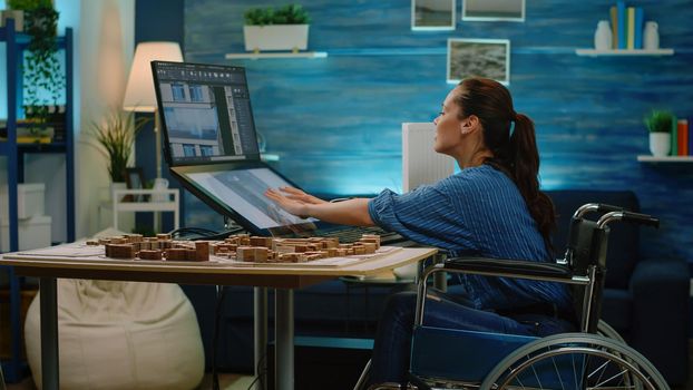 Engineer with disability using touch screen for building model at office. Invalid architect in wheelchair working on construction layout and blueprint plan while having maquette on desk