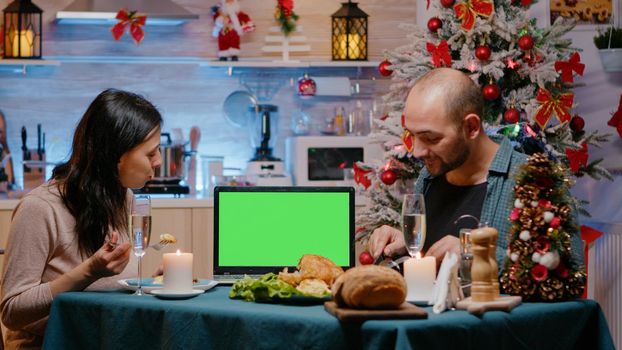Man and woman watching green screen on laptop enjoying festive dinner at christmas celebration. Couple looking at chroma key with mockup template and isolated background, celebrating holiday