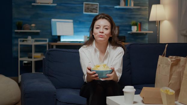 Portrait of caucasian woman eating chips from bowl while looking at camera, sitting on couch. Young adult with takeaway food on table in living room. Person with meal in delivery bags