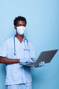 Black nurse holding laptop while looking at camera in studio. African american man working as medical assistant wearing face mask and gloves against covid 19 epidemic. Person with device