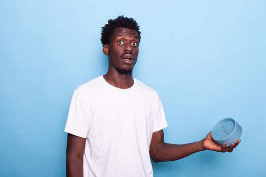 Young adult checking time on clock, being late in studio. Person showing hour and minute on watch, holding object with alarm and having worried expression on face. Punctual man in rush