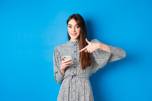 Stylish caucasian woman in dress, pointing at smartphone and smiling, showing online promo, standing on blue background.