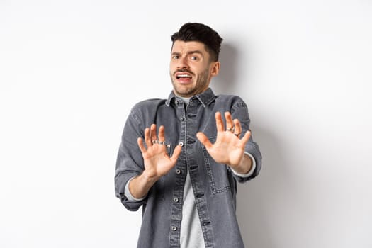 Stay away from me. Scared young man screaming and asking to stop, step back and look frightened, rejecting something bad, standing on white background.