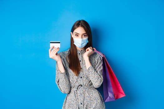 Covid-19, pandemic and lifestyle concept. Stylish woman wearing medical mask on shopping as preventive measure from coronavirus, showing plastic credit card and holding bags, blue background.