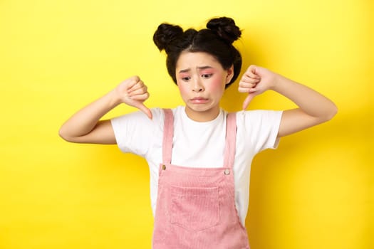 Asian beauty girl cringe from something disgusting, look away and showing thumbs-down to express dislike, negative emotions, standing in summer clothes on yellow background.