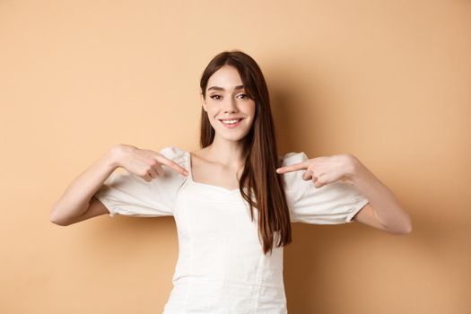 Smiling woman pointing at center, showing logo with happy face, standing in white dress on beige background.