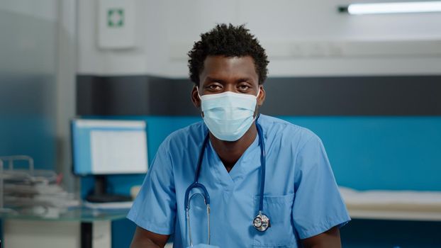 Portrait of man with nurse occupation looking at camera with face mask and uniform. Medical assistant sitting at desk in healthcare office with laptop and professional instruments