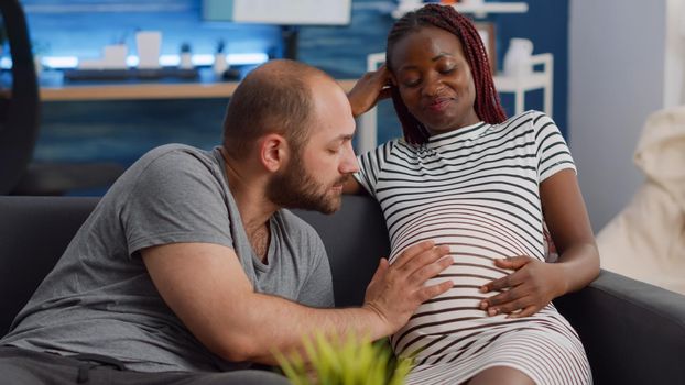 Married interracial couple expecting child bonding at home. Caucasian father talking to baby and touching belly of pregnant african american woman sitting on living room sofa.