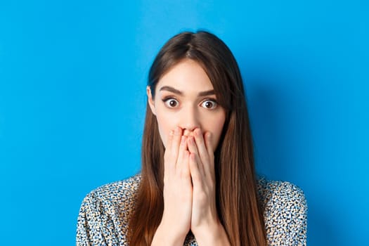 People emotions concept. Shocked young woman face, covering mouth with hands and gasping, look startled, standing on blue background.