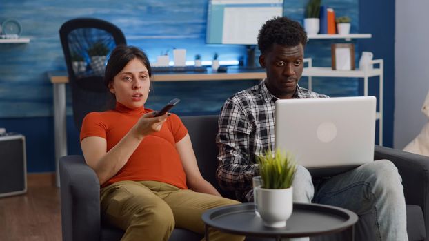 Interracial couple expecting child sitting together on couch. African american father of baby working on modern laptop while caucasian pregnant woman using TV remote control at home