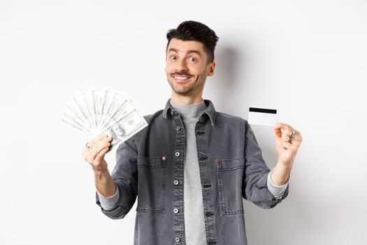Happy guy showing plastic credit card and dollar bills, giving choice, standing on white background.