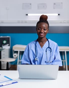 Portrait of african american nurse using laptop at white desk sitting in medical office. Black woman with assistant occupation wearing uniform and working at healthcare clinic