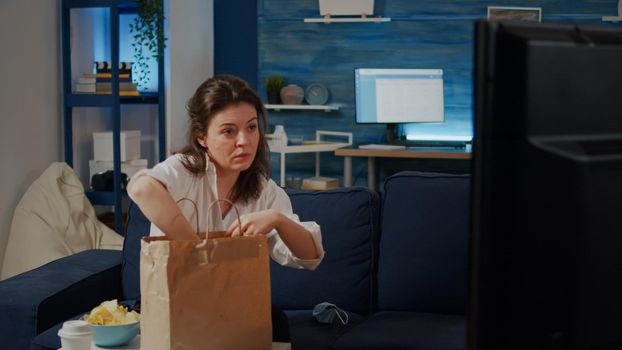 Young woman removing face mask after getting takeout food, placing bag on table in living room. Caucasian person preparing to eat unhealthy meal while watching television sitting on sofa