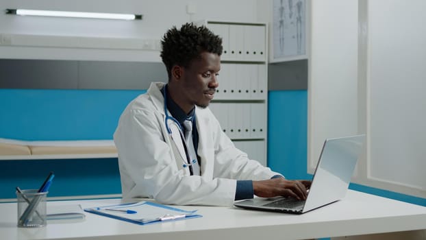 African american doctor using laptop while sitting at desk in hospital room. Black healthcare specialist working with technology for consultation in modern cabinet at medical facility