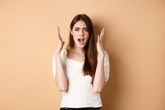 Frustarted woman shouting and shaking hands angry, looking mad, standing distressed on beige background.
