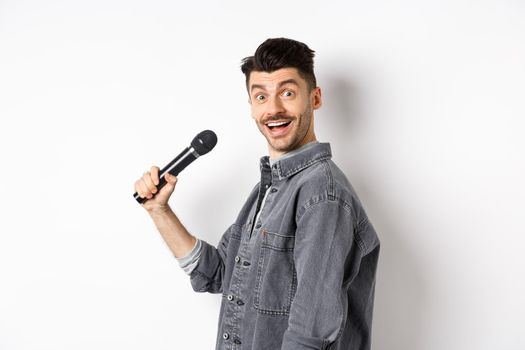 Profile of handsome smiling man holding mic, turn head at camera with excited face, singing karaoke and perform standup, standing on white background.