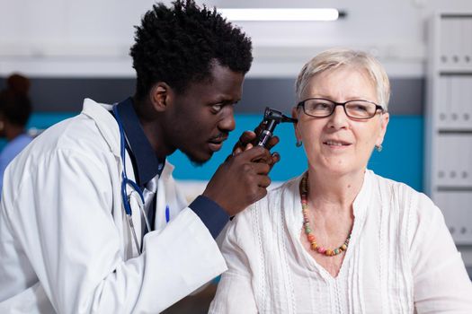 Black young doctor using otoscope on elder patient for ear consultation and examination. African american medic with metal tool for senior woman with healthcare problems in cabinet