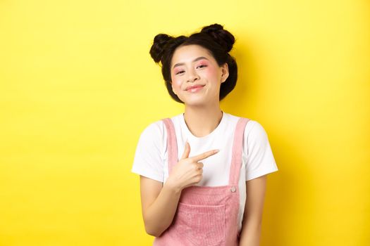 Stylish asian teen girl with romantic makeup, smiling cute and pointing finger right at logo, standing against yellow background.