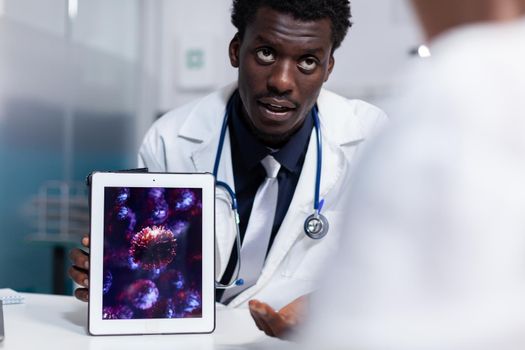 Doctor of african american ethnicity holding digital tablet with virus animation while sitting at desk. Black medic explaining disease bacteria and healthcare problems to old patient